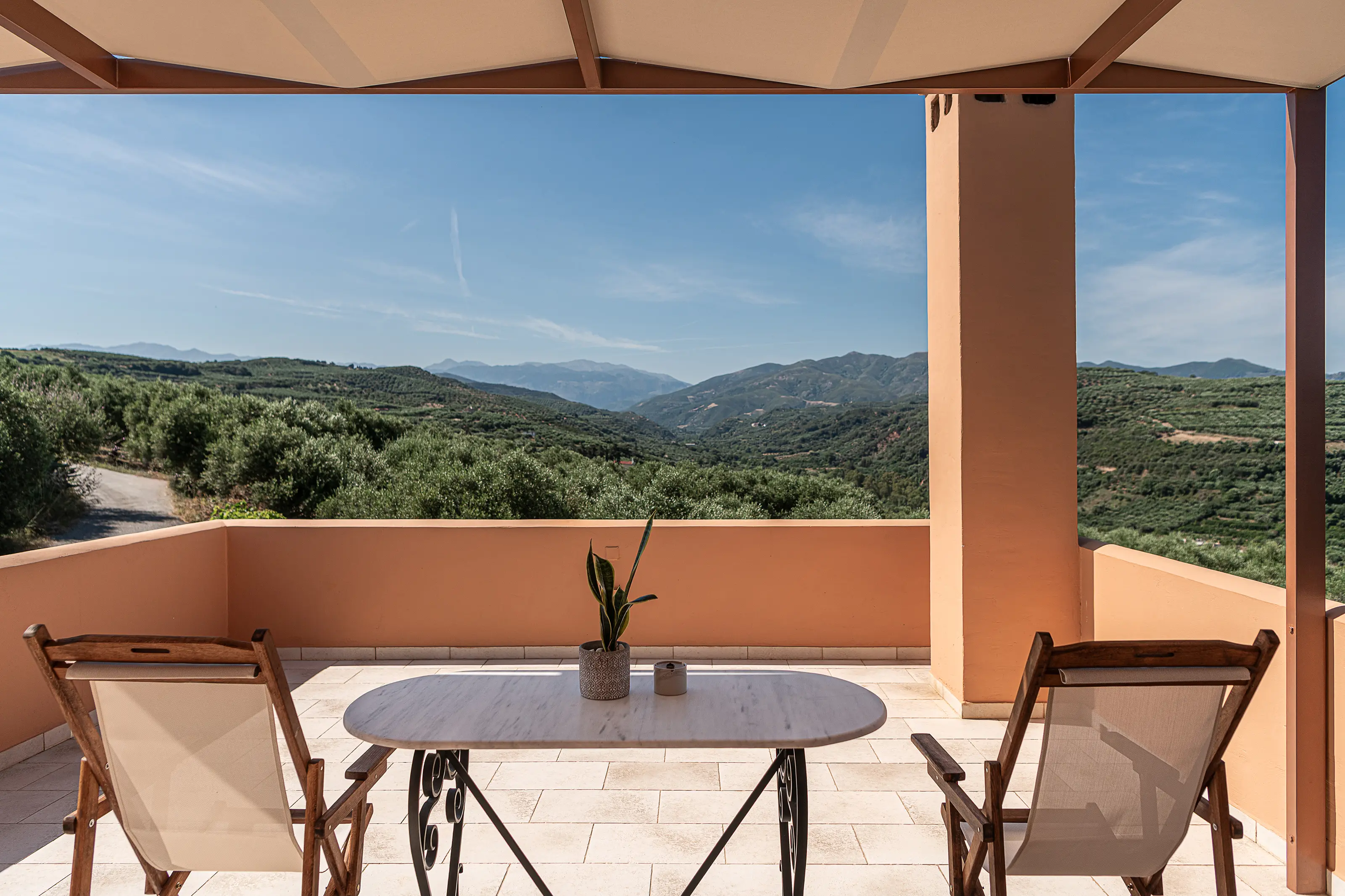 A shaded terrace with a table and two chairs, looking out over olive slopes to the mountains