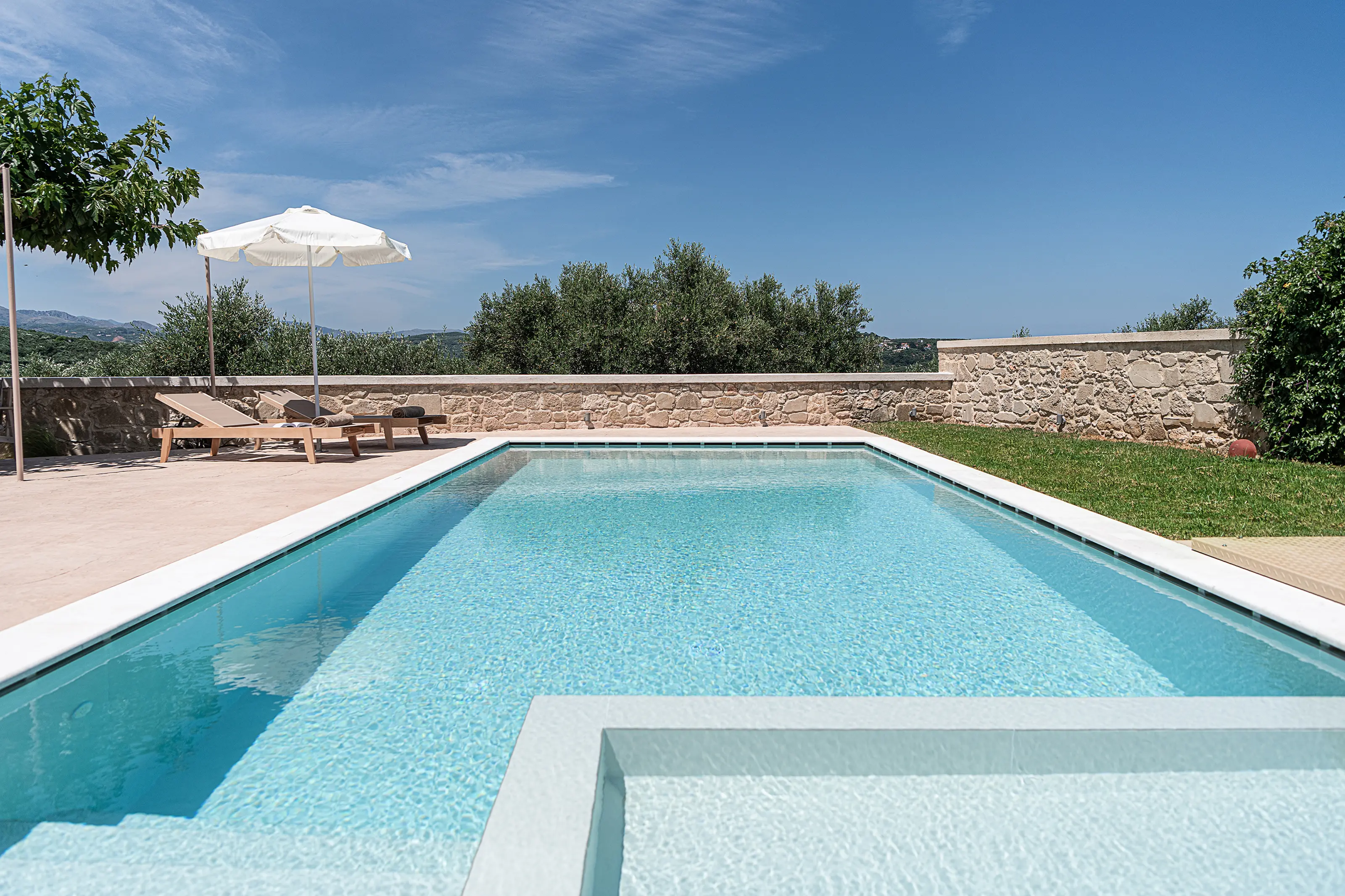 The pool from its near end: the hydromassage step in front, sunbeds and a parasol on the left, the olive groves and hills over the stone wall